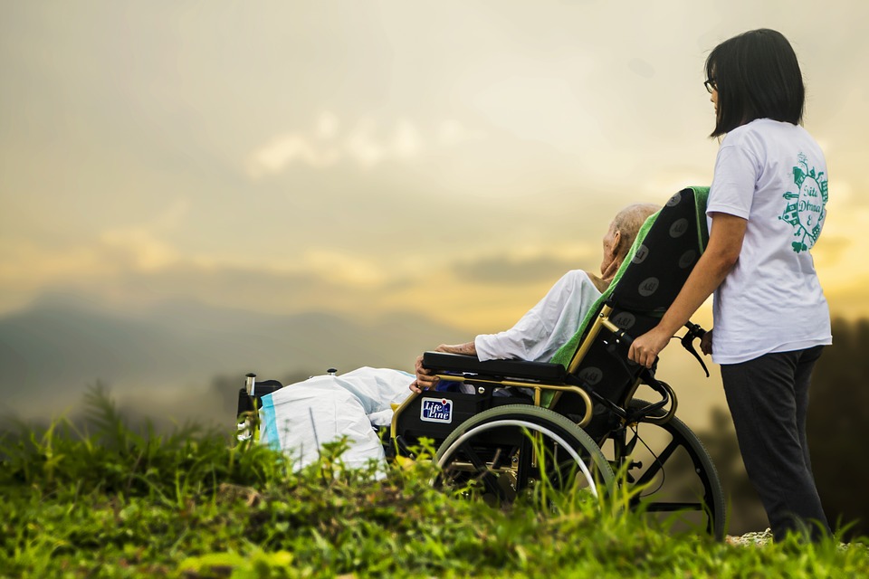 En äldre i en rullstol med ett vårdbiträde bakom sig